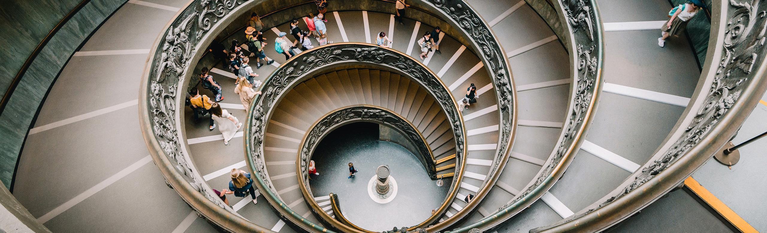 Top-down view of a monumental spiral staircase with ornate railing, filled with descending people.